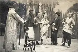 A man in traditional Chinese garb stands behind a camera, gazing upon a scene where a man carrying a cane and dressed in a bowler hat is chatting with a group of people in traditional Chinese clothing.