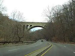 A four-lane full-access road with a low concrete median snakes through a wooded area and under a large stone arch bridge.