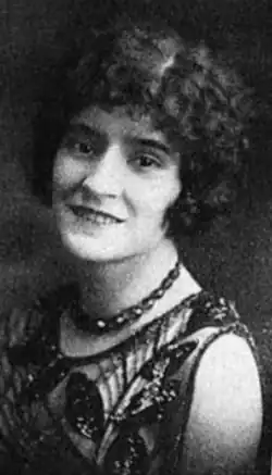 A smiling young woman with light skin and curly dark hair cut in a bob style, wearing a dark lace sleeveless dress and a short strand of chunky dark beads