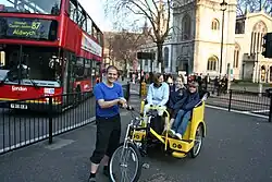 Three people sitting on the passenger seating of a pedicab while the driver stands next to the pedicab