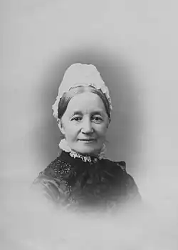 An older white woman, wearing a white cap and a high-collared garment. She is smiling slightly, and her hair is center parted and dressed back.