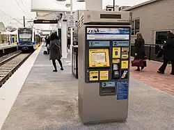 A silver and blue ticket vending machine on a station platform with payment options and digital screen visible.