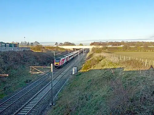 A photograph of an active railway line in a cutting passing underneath a motorway. The cutting's embankments on each side are grassy and steep.