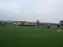 A sports centre with two buildings, the left a modern square building with a wood upper floor above a glass lower floor and the right metal and glass, behind green playing fields