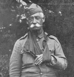 A black and white photo of a man with an impressive white mustache. His arm is in a sling and he is wearing a military cap.