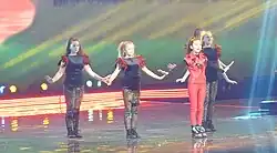Manw in red standing on stage with four dancers in black standing diagonally behind her
