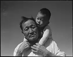 A black and white photo of an elderly, Japanese grandfather and his young Japanese grandson, on his shoulders at Manzanar Relocation Center, part of the photos that were impounded during the war