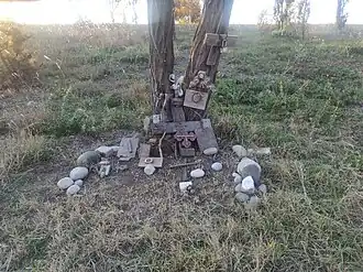 A rustic, handmade memorial at the base of a tree in a grassy field. It consists of an assemblage of weathered wood pieces, some forming crosses, and various stones.