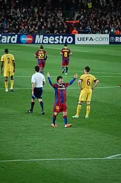 Male football players representing FC Barcelona (red and blue stripes) and Arsenal F.C. (yellow) wearing different colour outfits, while the referee is in a white T-shirt.