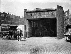 A horse and cart stand in a street outside a narrow building with large open doors and a dark interior. A sign above the doors says "Metropolitan Railway Goods Depot".