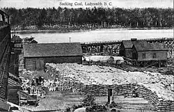 a black and white photo of an industrial workplace on the shore. There is a large wooden building with chutes to the left, huge piles of white sacks in the foreground, and a trestle bridge and sheds on posts in the background