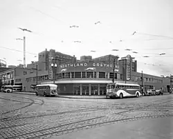 Black and white photo of corner building on a semi deserted street with round facade, sign saying Northland Greyhound, and two vertical signs reading Greyhound, surrounded by buses, Pantages Vaudeville and other buildings visible in rear