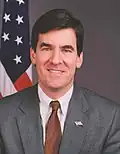 White male with dark hair in a gray suit with an American flag in the background