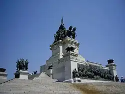 Photograph of a white stone steps leading up to a large, altar-like monument in white marble with bronze sculptural decorations that include bronze braziers at the corners, a bronze frieze in high relief at the base and bronze figures surrounding a chariot on a high, white marble plinth in the center