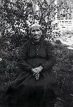 Mother Solomon wearing a patterned dress and sitting in front of a bush with her hands in her lap.