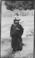 Rumbur Valley, Chitral, NWFP, Pakistan Kalash girl in Rumbur. The girl is dressed in a dark gown and carries jewellery: a ring, a necklace and a headdress (kupa) covered with shells (kauri or cowri). On top of the headdress there is a tassle (probably red) and a second one, smaller, is visible above her forehead. Her shoes are also clearly visible. 1929 approx.