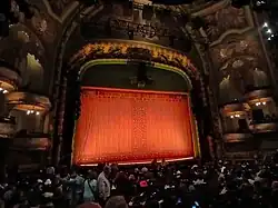 The proscenium arch as viewed from the rear of the orchestra level's left side. There is a red curtain within the opening. The arch is ornately decorated and is flanked by pairs of double-height staggered box seats.