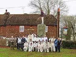 Two cricket teams plus umpires, scorers and groundsman assemble at granite monument on side of cricket pitch by the bat and ball pub for a group photo