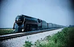 A black streamlined steam locomotive pulling a rake long line of passenger cars in the rural countryside.