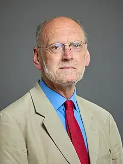 close-up of Nigel Biggar wearing a tan-coloured blazer and light blue shirt with a red patterned tie, looking directly at camera with serious gaze