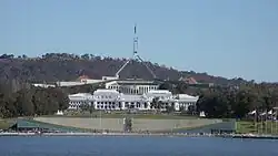 Bright white classical building in front of larger, modernistic beige, gray, and green building. Two metal spires stretch from the top of the larger building in a trangular shape; near the top, before the two connect, they bend straight up, supporting a flagpole and flag.
