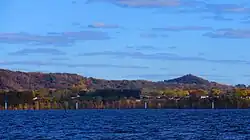A landscape photo of Onalaska Wisconsin taken in the fall from Lake Onalaka shows a sunny sky of blue above with clouds and the cool blue waters of Lake Onalaska below in the foreground. Sandwiched between is a horizontal ribbon that shows the city elevated 100 feet above the water and tree covered bluffs in the not to distant background. A few houses along highway 35 are barely visible.