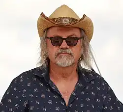 Ondřej Hejma wearing a dark blue patterned shirt, straw cowboy hat, and black sunglasses, looking directly at camera
