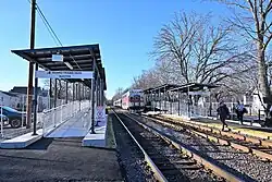 Short sections of high-level railway platforms with ramps, with a train departing and passengers crossing the tracks.