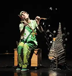 Someone on the street sitting on a bin playing a saw with a bow and with it between his legs.