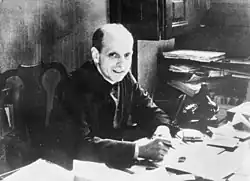 Black and white photograph of a bald white man in a black cassock looking at the viewer, smiling and holding a pen, sitting at a busy desk with documents and a black telephone.