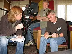A photograph of four men in a room, three of them sitting down, all surrounding a black videocamera on a black tripod with windows in the background