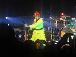 In the middle of a stage, a man wearing a yellow suit and a red hat plays a guitar. In the background is a similarly dressed man playing drums. A crowd is visible in the foreground.