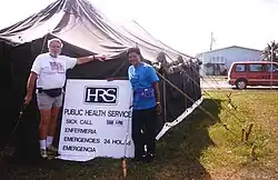Two people standing outside a large black tent, beside a sign reading 'HRS public health services. Sick call Enfermeria 8am–6pm. Emergencies Emergencia 24 hours.'