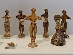 Six clay figurines, humanoid, with their fists held to their chests. Most are male; one female figurine has a large, bell-shaped skirt.