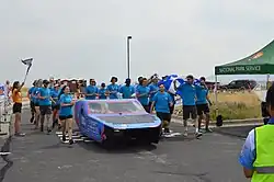 The 2 Person Solar Vehicle Built by Students at Polytehnique Montreal Crosses the Electrek American Solar Challenge 2024 finish line as the checkered flag waves and team members run behind the car cheering for their accomplishment.