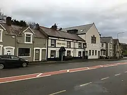 Main A470 road in foreground of black tarmac with red central stripe having residential buildings, pub and Memorial Hall stretched behind into distance away from camera position