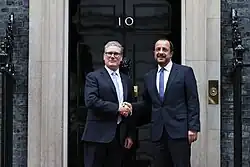 Two men dressed in dark suits shake hands in front of the black door to 10 Downing Street.
