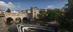 A three arch stone bridge with buildings on it, over water. Below the bridge is a three step weir and pleasure boat.