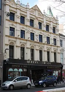 An ornate Victorian 4-storey building, painted pastel yellow, featuring muted copper spires on the roof, a small white clockface in the centre of the 2nd floor, and ground floor shopping units framed in black, either side of the arched entrance to an arcade. On the third floor is an Art-Deco-influenced monogram with the letters "A.R.".