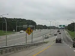View of a three-lane collector/distributor road of a freeway; several green signs are visible in the distance. At the center of the image is a four-lane divided freeway that is separated from the distributor road by concrete barriers.