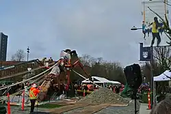 Action shot of people, wearing orange and yellow construction clothing, working to raise the Reconciliation Pole at UBC