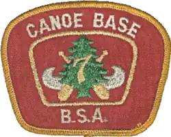 Patch with a red background and a gold border; the text Canoe Base BSA in the border; in the center a silver canoe superimposed with crossed paddles, a green pine tree and the number 7 in gold