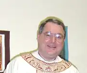 A bespectacled man wearing a white chasuble with beige and burgundy ornamentation around the neckline