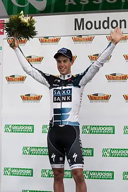 A man in his early twenties wearing a white and black cycling jersey with blue trim and a matching cap. He is holding his arms up in a posture of victory, and holds a bouquet in his right hand.