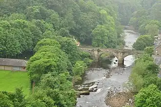 An elevated view looking down on a stone bridge crossing a river showing three arches, and either bank of the river lined with trees