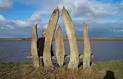 View from the rear of a seat made from 5 vertical rough-hewn planks of wood, with a person sitting on the seat and looking out over a wide river