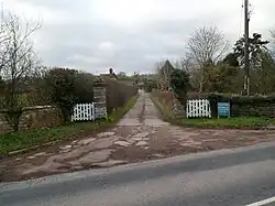A road entrance to a building consisting of white-painted wooden gates, facebrick walling in a green landscape which prominently features various tree species.