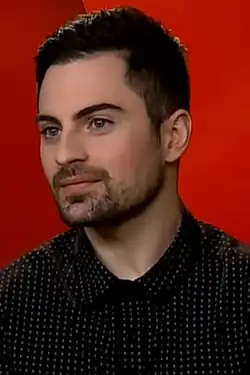 close-up of Rodolfo Valente in front of a bright red background, wearing a a dark collared t-shirt with dots, smiling slightly and looking right of camera