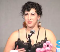 A woman with curly black hair and large disc earrings with scorpions on them speaks into a microphone in front of a gray wall.