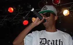 A black male with sunglasses, a white shirt with text “Death,” and a green and white snapback speaking into a microphone